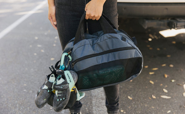 Person holding an OSCA that has some climbing shoes attached via a carabiner to the shoulder strap attachment point.